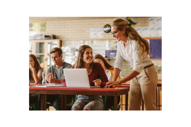 sala szkolna, 4 młode osoby siedzą w ławkach, nauczycielka pochyla się nad uczennicą na pierwszym planie