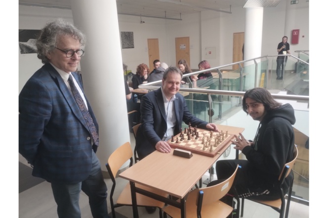 2 osoby siedzą przy stoliku z szachami, mężczyzna stoi obok