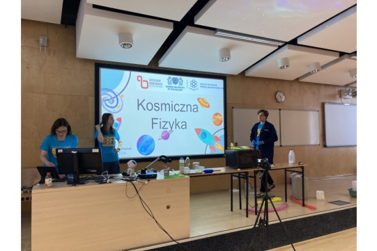 trzy osoby na scenie, w tle tablica z napisem "Kosmiczna fizyka"