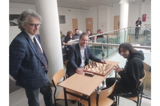 2 osoby siedzą przy stoliku z szachami, mężczyzna stoi obok