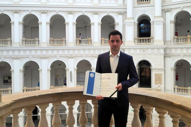 The photo shows MSc Jacek Szkopek dressed in a dark suit and white shirt holding a diploma in his hand, which he obtained winning the third place in the 9th Siemens prize competition. The man is standing on the terrace of the courtyard.