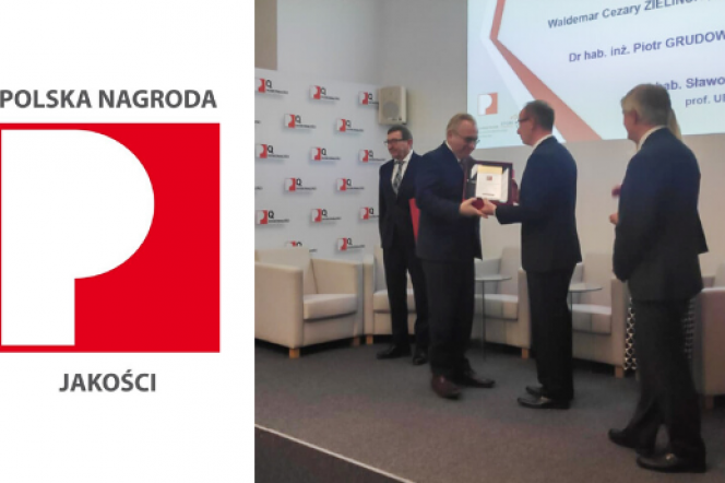 The picture shows the logo of the Polish Quality Award, i.e. the white letter P on a red background. Apart from the logo, there are four men dressed in dark suits and one woman standing behind the man PhD eng. Piotr Grudowski is receiving the Polish Quality Award.