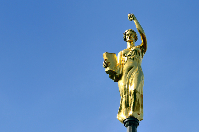 Zdjęcie przedstawia rzeźbę Alegorii Nauki w kolorze złotym, która wieńczy wieżę zegarową gmachu głównego Politechniki Gdańskiej na tle niebieskiego nieba
