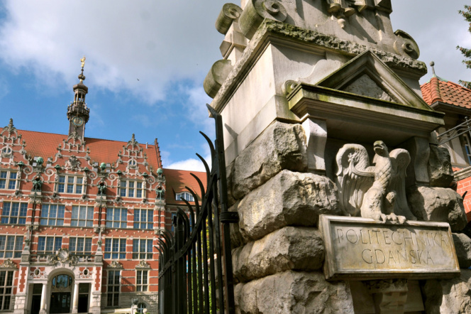 Na zdjęciu widoczny fragment bramy głównej z napisem Politechnika Gdańska z orłem. W tle gmach główny Politechniki Gdańskiej