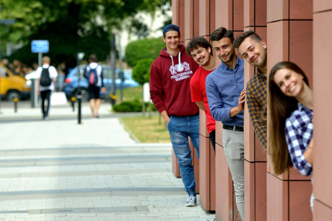 Zdjęcie przedstawia grupę studentów wyglądających zza filarów budynku