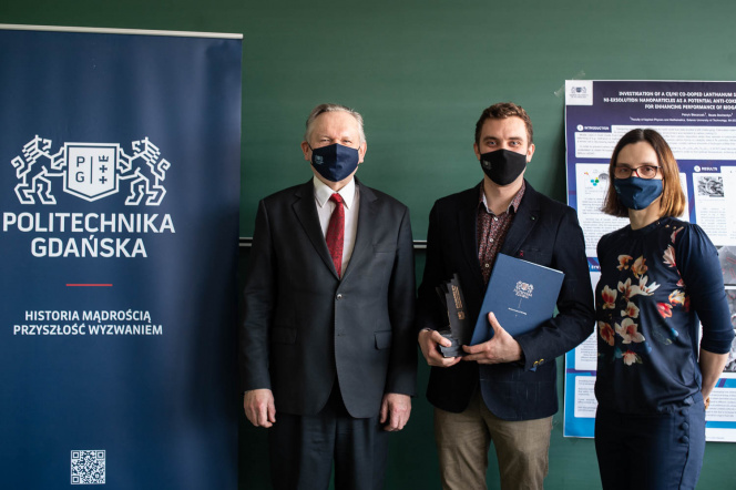 The photo shows the laureate of M.Sc. Patryk Blaszczak in the company of prof. dr hab. Józef Sienkiewicz and the supervisor, dr hab. Eng. Beata Bochentyn. Everyone is standing next to each other against the background of a green wall and a navy blue banner with the Gdańsk University of Technology. All are dressed in dark.