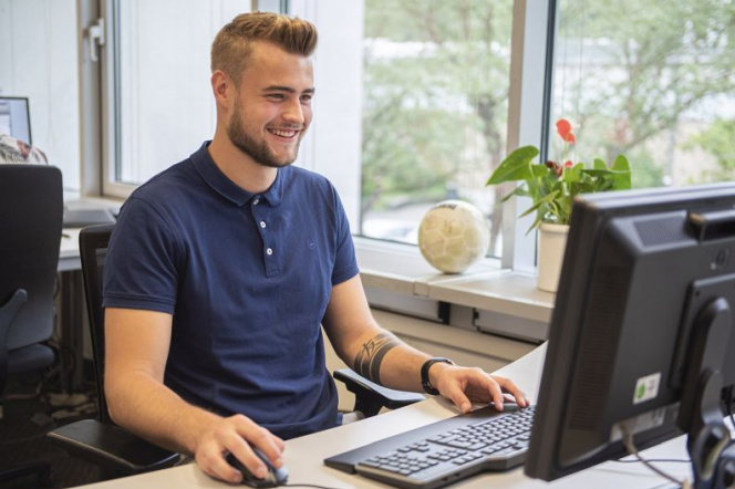 Na zdjęciu znajduje się mężczyzna ubrany w granatową koszulkę. Siedzi przy biurku i pracuje na komputerze.