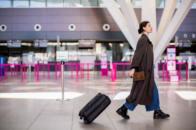 The picture shows a woman wearing a brown long coat. While walking at the airport she is dragging a suitcase behind her. 