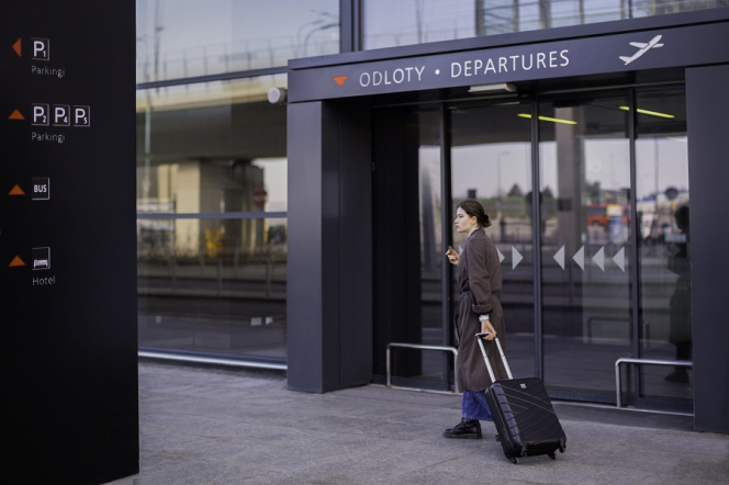 Na zdjęciu na  tle drzwi do lotniska znajduje się kobieta w brązowym płaszczu z walizką.