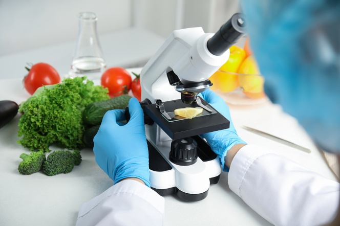 Dłonie naukowca przy mikroskopie, wokoło warzywa i owoce.