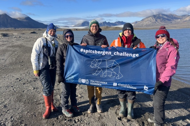 Na tle krajobrazu Spitsbergenu pięcioro studentów. Trzymają granatowy baner z logotypem wyprawy Spitsbergen Challenge.