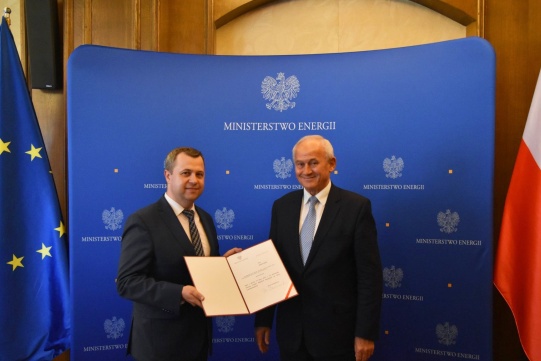 Na zdjęciu znajduje się dwóch mężczyzn: absolwent Politechniki Gdańskiej dr inż. Rafał Gawin oraz minister energii Krzysztof Tchórzewski. Obaj mężczyźni stoją się koło siebie, ubrani są w ciemne garnitury i jasne koszule. Tło zdjęcia jest niebieskie po jednym boku znajduje się flaga Unii Europejskiej, po drugim boku znajduje się flaga Polski.