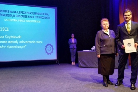 In the foreground picture there is a young man dressed in a dark suit and a woman dressed in a dark jacket, a light shirt and a black skirt. In the background there is a young woman dressed in a dark grey suit. 