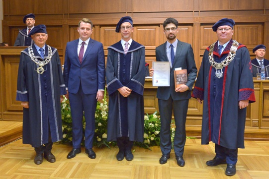Na zdjęciu się pięciu mężczyzn, w tym prof. Krzysztof Wilde Rektor PG oraz absolwent inż. Cezary Swinarski.  Dwóch mężczyzn ubranych jest w garnitury, trzech w togi. Mężczyźni stoją w auli.