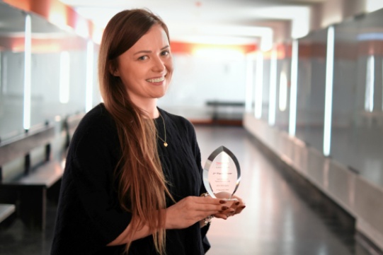 Na zdjęciu znajduje się mgr inż. Dorota Garwolińska trzymająca w rękach zdobytą nagrodę statuetkę. Kobieta o brązowych długich włosach ubrana jest w czarną zwiewną sukienkę, pięknie się uśmiecha.
