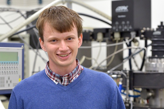 Na zdjęciu znajduje się Krzysztof Pyrchla. Mężczyzna o jasnych włosach ubrany w niebieski sweter i koszulę w kratkę stoi na tle różnych aparatur.