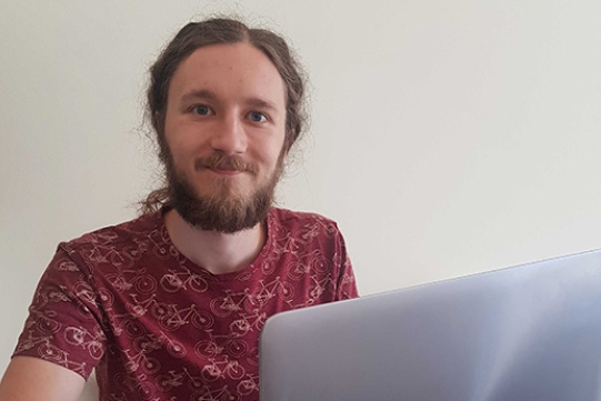 Na zdjęciu znajduje się dr inż. Miłosz Wieczór ubrany w bordową koszulkę w żółte rowery. Włosy ma upięte w kitkę, ma brodę.