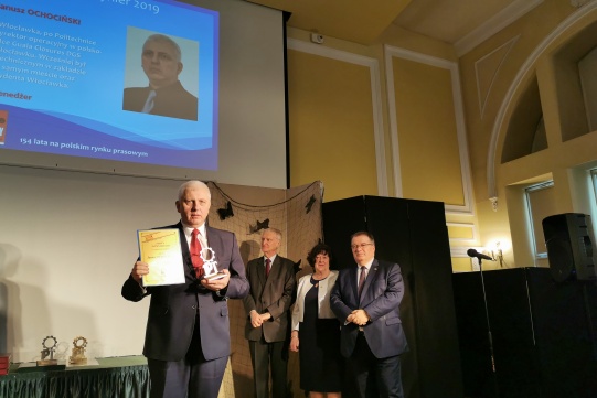 Na zdjęciu na pierwszym planie znajduje się mgr inż. Janusz Ochociński ubrany w ciemny garnitur, trzymający w rękach zdobytą statuetkę Złotego Inżyniera oraz dyplom. Na drugim planie znajduje się dwóch mężczyzn ubranych w ciemne garnitury oraz kobieta ubrana w kremową marynarkę i czarną sukienkę. 