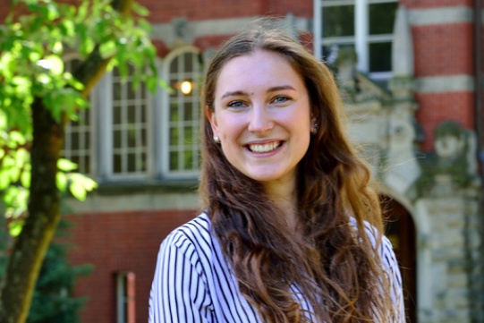 Na zdjęciu znajduje się mgr inż. Justyna Górska uśmiechnięta brunetka ubrana w koszulę w biało granatowe pionowe paski. Stoi na tle budynku z czerwonej cegły.