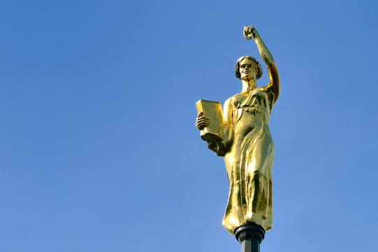 Zdjęcie przedstawia rzeźbę Alegorii Nauki w kolorze złotym, która wieńczy wieżę zegarową gmachu głównego Politechniki Gdańskiej na tle niebieskiego nieba