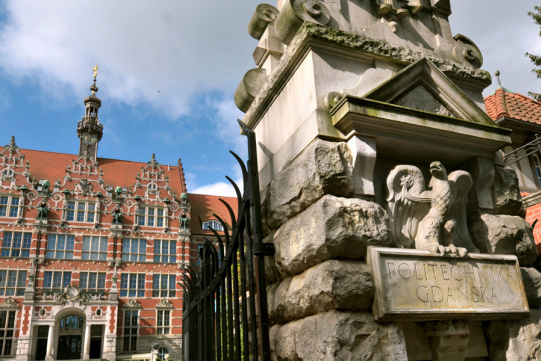 Na zdjęciu widoczny fragment bramy głównej z napisem Politechnika Gdańska z orłem. W tle gmach główny Politechniki Gdańskiej