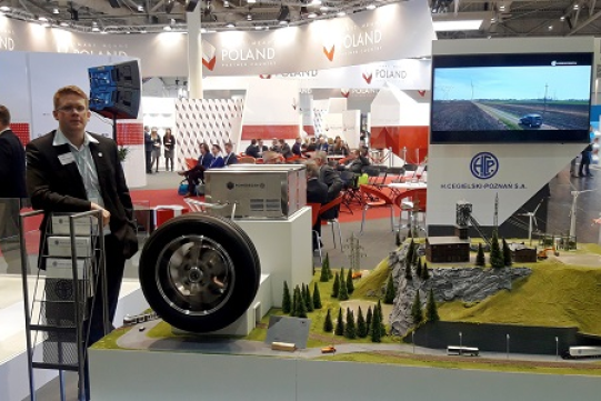 Na zdjęciu znajduje się dr inż. Patrick Strankowski laureat konkursu o Nagrodę Simensa ubrany w ciemny garnitur i jasną koszulę. Stoi w pomieszczeniu targowym, gdzie znajdują się stoiska wystawiennicze.