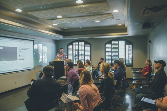 poglądowe zdjęcie przedstawiające grupę ludzi siedzących na szkoleniu. Ich uwaga skupiona jest na prezentacji wyświetlanej przez prowadzącego na rzutniku