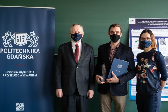 The photo shows the laureate of M.Sc. Patryk Blaszczak in the company of prof. dr hab. Józef Sienkiewicz and the supervisor, dr hab. Eng. Beata Bochentyn. Everyone is standing next to each other against the background of a green wall and a navy blue banner with the Gdańsk University of Technology. All are dressed in dark.