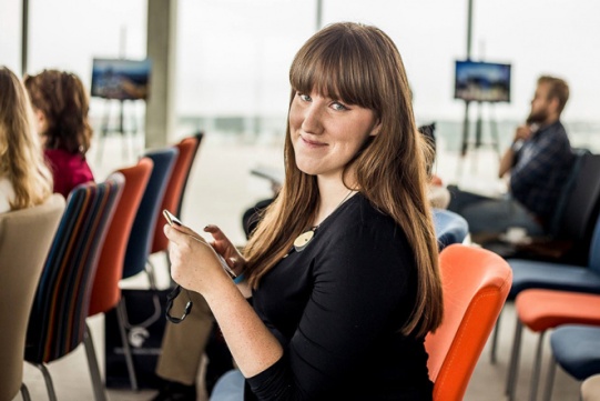 The photo shows Dr. Eng. arch. Hanna Obracht-Prondzyńska from the Faculty of Architecture. The woman has long brown hair with fringe and is wearing a dark blouse with short sleeves. He is standing in the hall against the background of colorful chairs