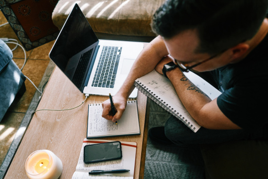 zdjęcie przedstawia studenta siedzącego przed laptopem i notującego w zeszycie