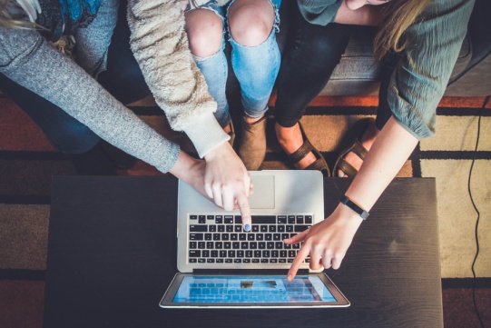 na stole leży laptop, a w okół niego siedzą 3 studentki, które coś wskazują na monitorze
