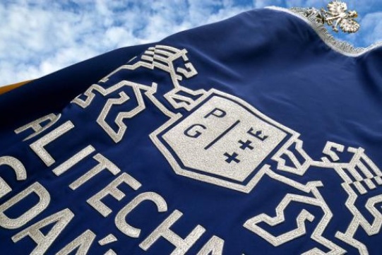  The photo shows a navy blue flag with the white logo of the Gdańsk University of Technology against the sky.