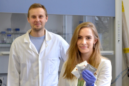 Na zdjęciu znajduje się mgr inż. Zuzanna Zarach, która otrzymała nagrodę oraz promotor dr inż. Mariusz Szkoda. Obydwoje ubrani są w białe kitle. Obydwoje mają blond włosy, kobieta o długości za ramiona.