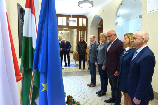 Pięcioro ubranych elegancko osób stoi na baczność w szeregu, przed tablicą pamiątkową.