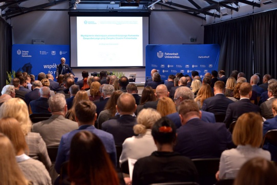 Sala konferencyjna wypełniona uczestnikami wykładu. W drugim planie biały ekran, mównica. Przy mównicy ubrany w garnitur mężczyzna