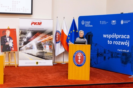 Mównica na auli uniwersyteckiej. Na mównicy logotyp GUMed. Przy mównicy kobieta. W drugim planie rozmaite roll-upy i ścianki promocyjne.