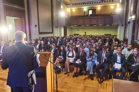 Mężczyzna przemawiający przy mównicy. W drugim planie aula wypełniona elegancko ubranymi osobami.