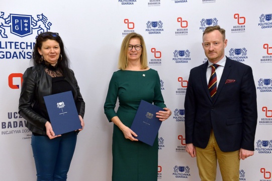 Na zdjęciu trzy osoby - dwie kobiety i meżczyzna. Kobiety trzymają niebieskie teczki z umowami.