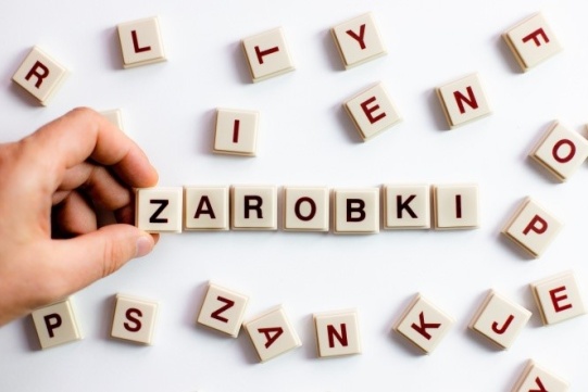 Na zdjęciu widoczna jest dłoń układająca wyraz ZAROBKI z porozrzucanych taliczek z lierami 