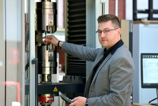 Na zdjęciu znajduje się mężczyzna ubrany w szarą marynarkę i ciemny golf