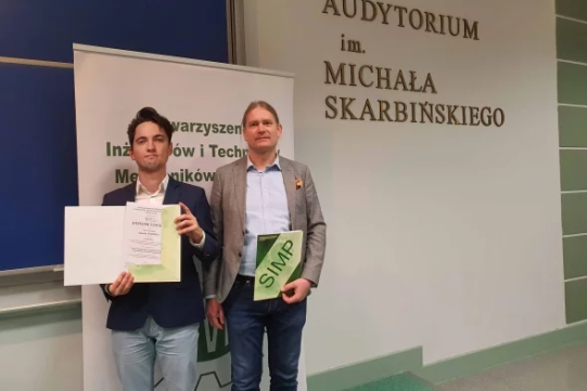 Na zdjęciu znajduje się dwóch mężczyzn stojących na tle białych banerów