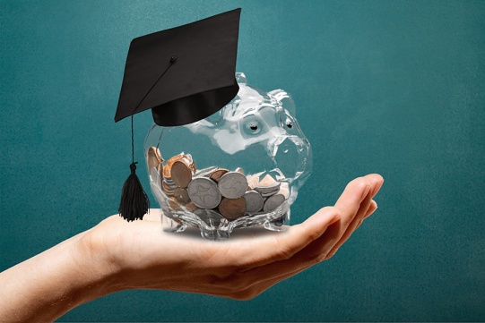 dłoń na której lezy skarbonka świnka przeźroczysta z monetami, skarbonka na głowie ma czarny biret, tło w ciemnozielonym odcieniu