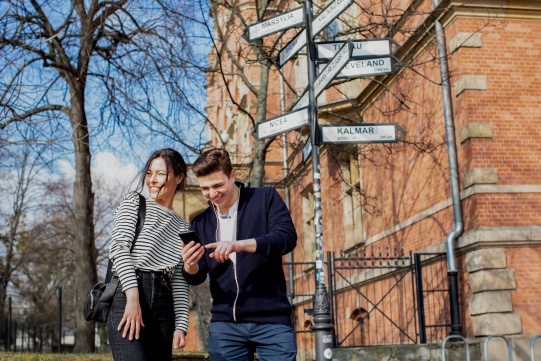two smailing students on the campus, standing next to the signs with the names of different European cities