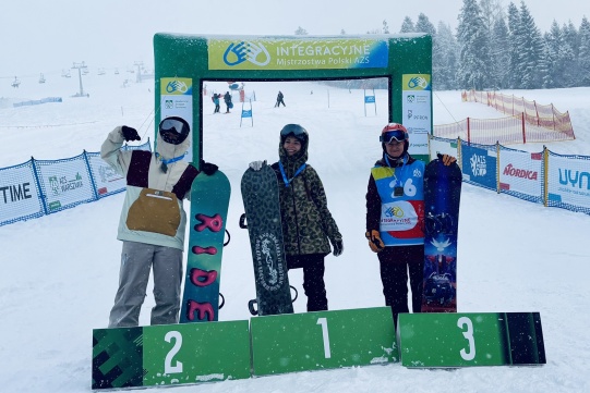 Trójka snowboardzistów na podium zawodów Integracyjnych Mistrzostw Polski AZS na tle ośnieżonego stoku narciarskiego.
