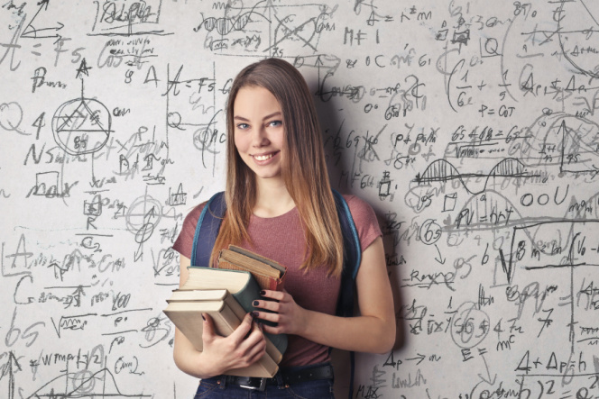 Zdjęcie przedstawia uśmiechniętą studentkę w rozpuszczonych włosach, która w ręku trzyma 4 książki. Studentka stoi na tle białej tablicy z licznymi wzorami matematycznymi.