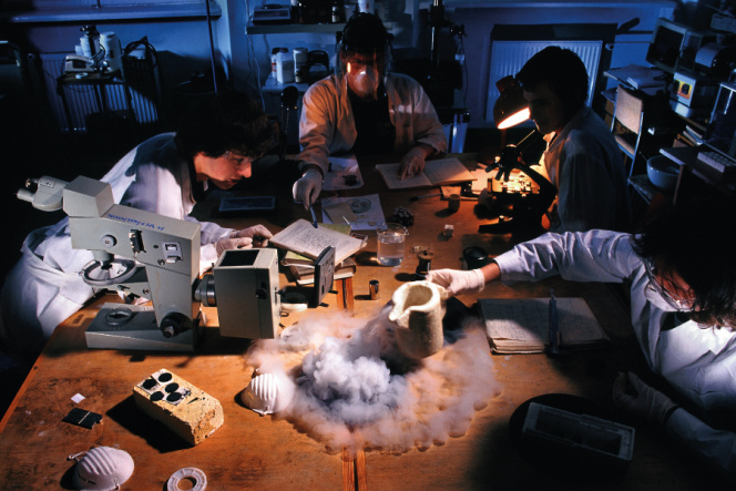 Zdjęcie przedstawie 4 studentów w białych fartuchach laboratoryjnych, którzy siedzą wokół stołu i robią doświadczenia chemiczne. Świadło w pomieszczeniu jest przyciemnione. Na stole znajdują się ksiązki, notatki, mikroskop, zlewka z wodą.