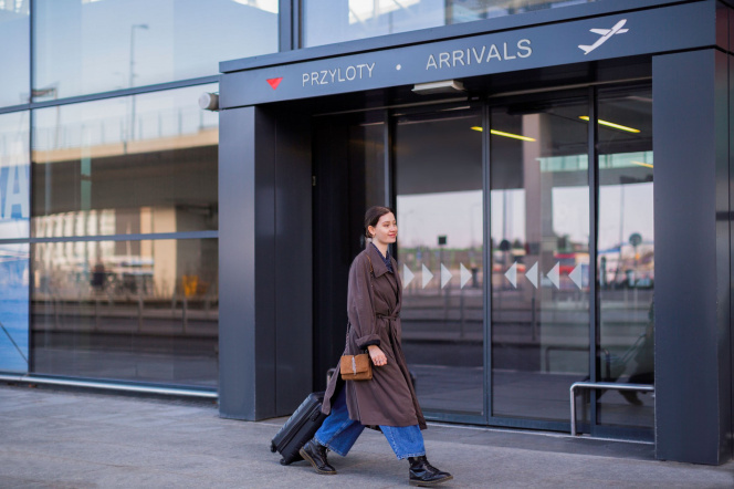 Na zdjęciu widać młodą kobietę z walizką przed wejściem na lotnisko. 
