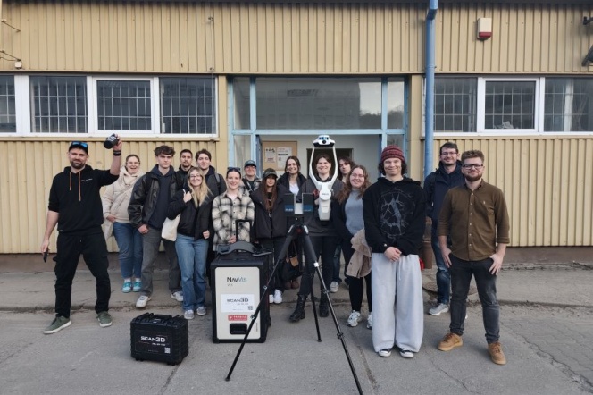 Grupa kilkunastu studenci Geodezji i Kartografiistoi przed wejściem do budynku o żółtej elewacji. W centrum ustawiono sprzęt pomiarowy na statywie oraz walizki z urządzeniami, sugerujące zajęcia terenowe lub prezentację technologii.