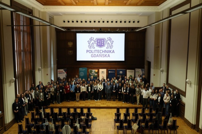 Zdjęcie grupowe uczestników konferencji „Transport i Urbanistyka 2026” wykonane w auli Politechniki Gdańskiej. Kilkadziesiąt osób stoi na scenie na tle dużego ekranu z logotypem uczelni oraz roll-upów organizatorów i partnerów wydarzenia.