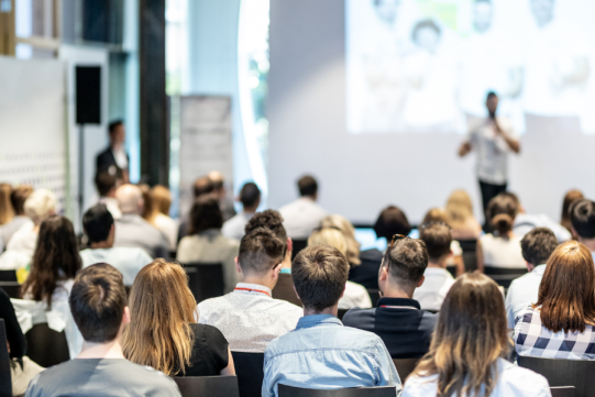 Ludzie siedzący na krzesłach słuchają mężczyzny, prezentującego slajdy na ekranie. 